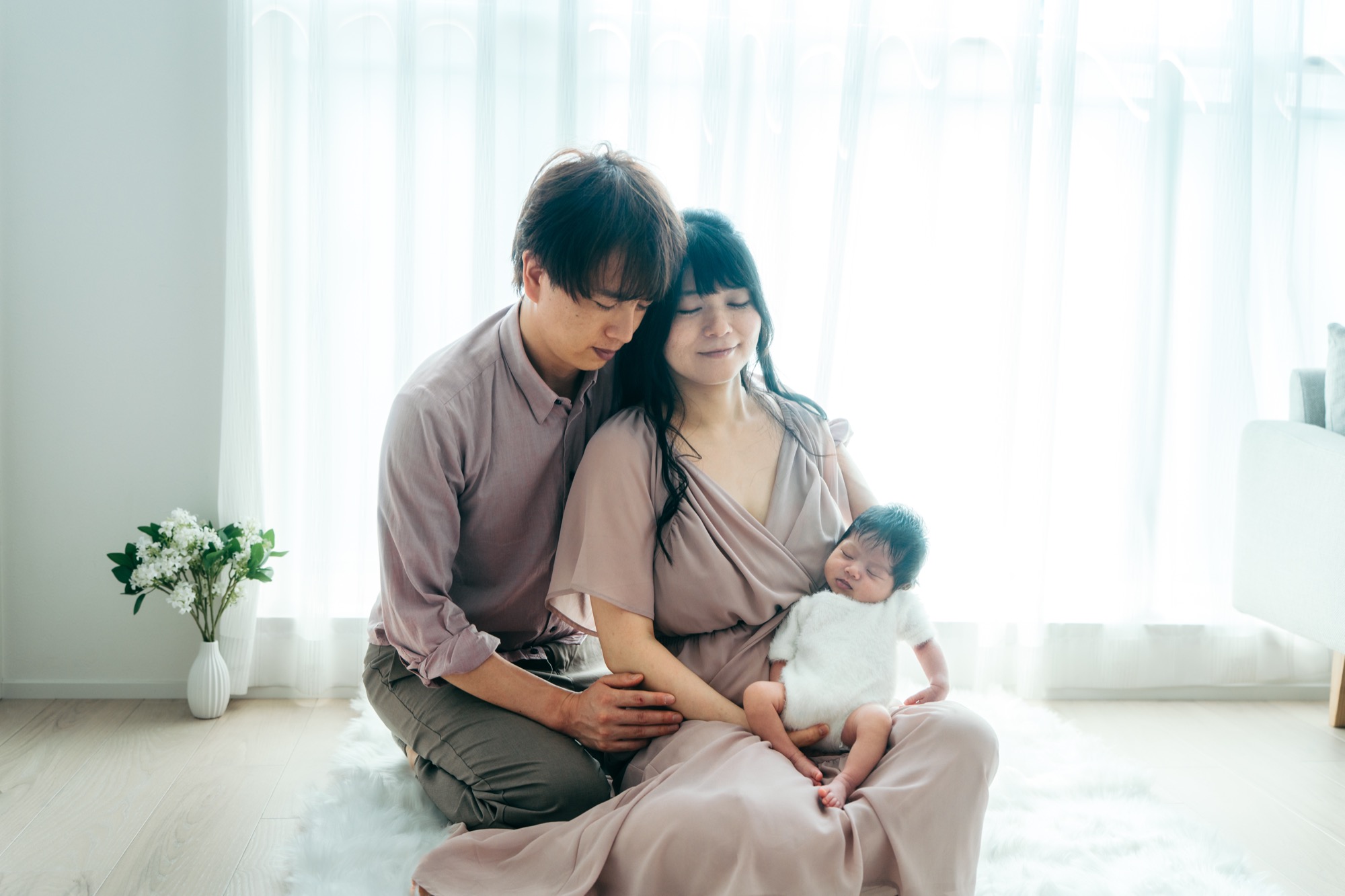 マタニティフォトと同じ座りポーズの産後比較カット：窓辺でママの膝に眠る赤ちゃんと寄り添うパパのニューボーンフォト