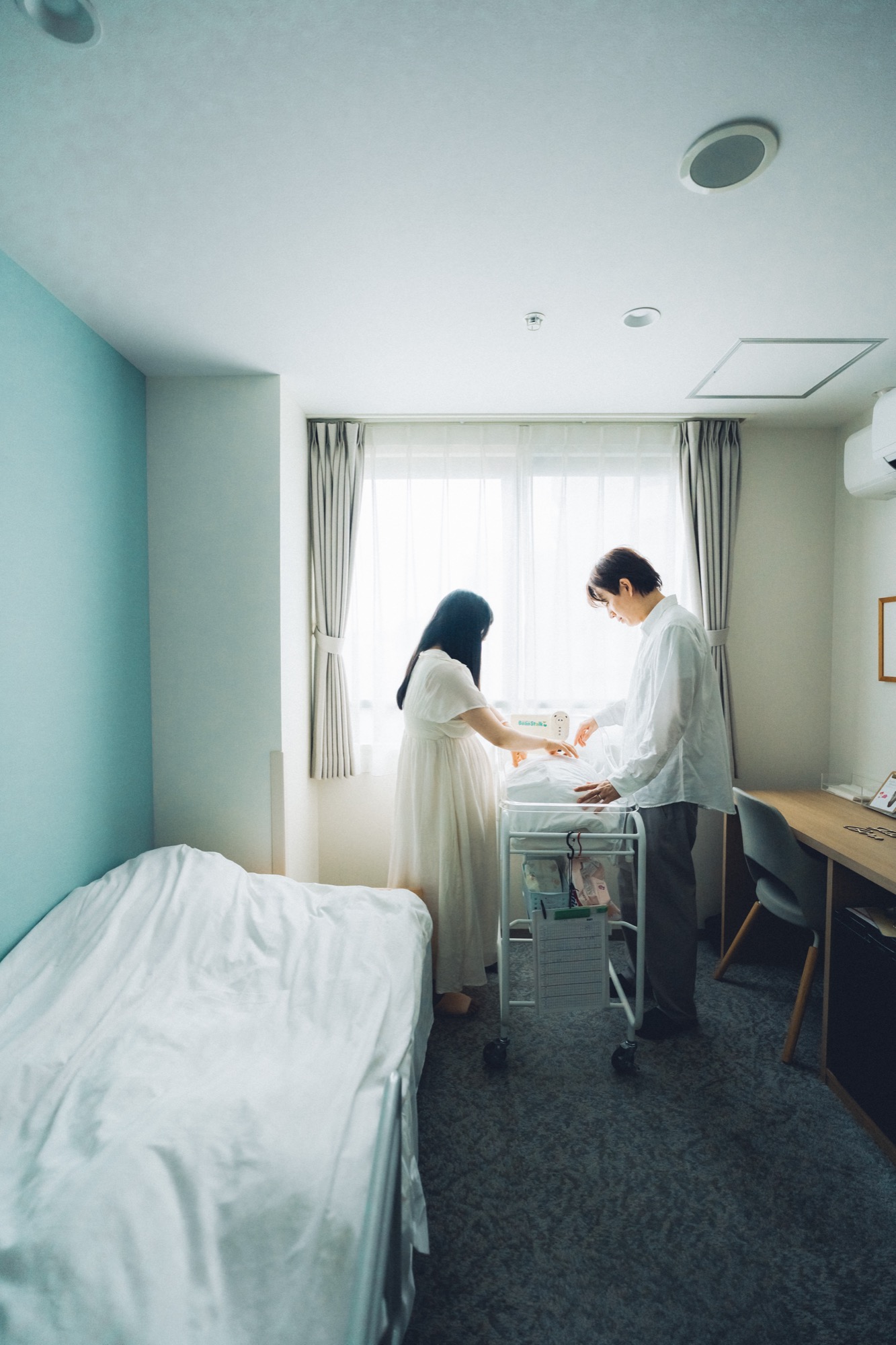 産院の病室でベビーコットの赤ちゃんを見守るご夫婦のニューボーンフォト