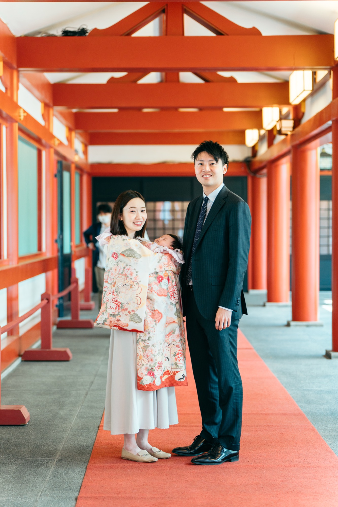 祝い着を羽織ったママとパパが、朱の回廊の赤い絨毯の上で並ぶお宮参りの記念写真