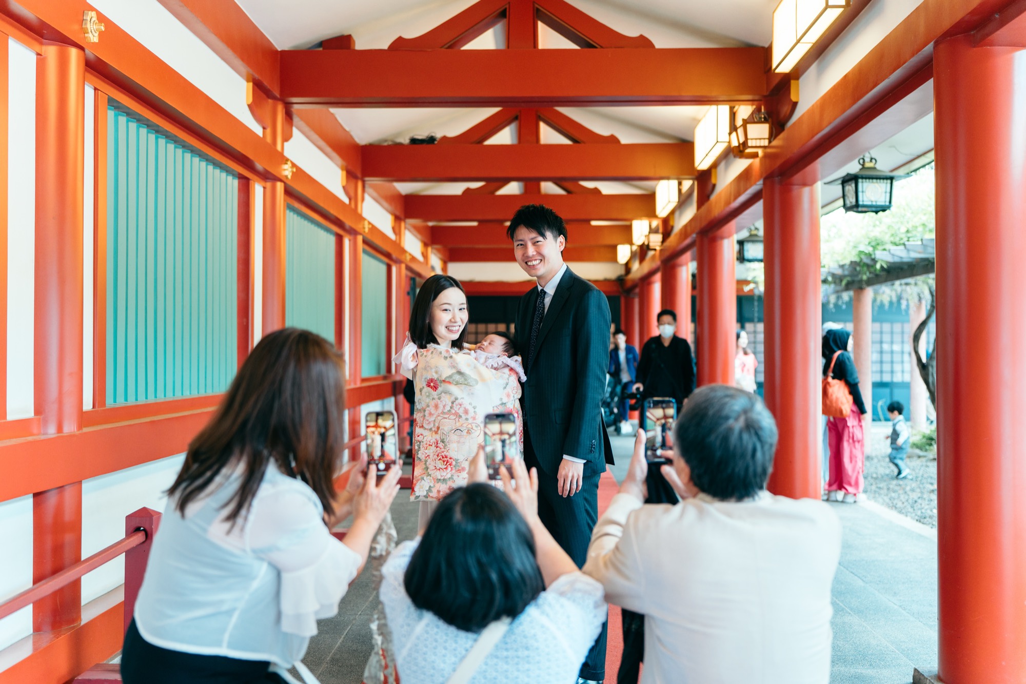 回廊で、お宮参りのご夫婦をスマホで撮影する祖父母たち。和やかな家族のひととき