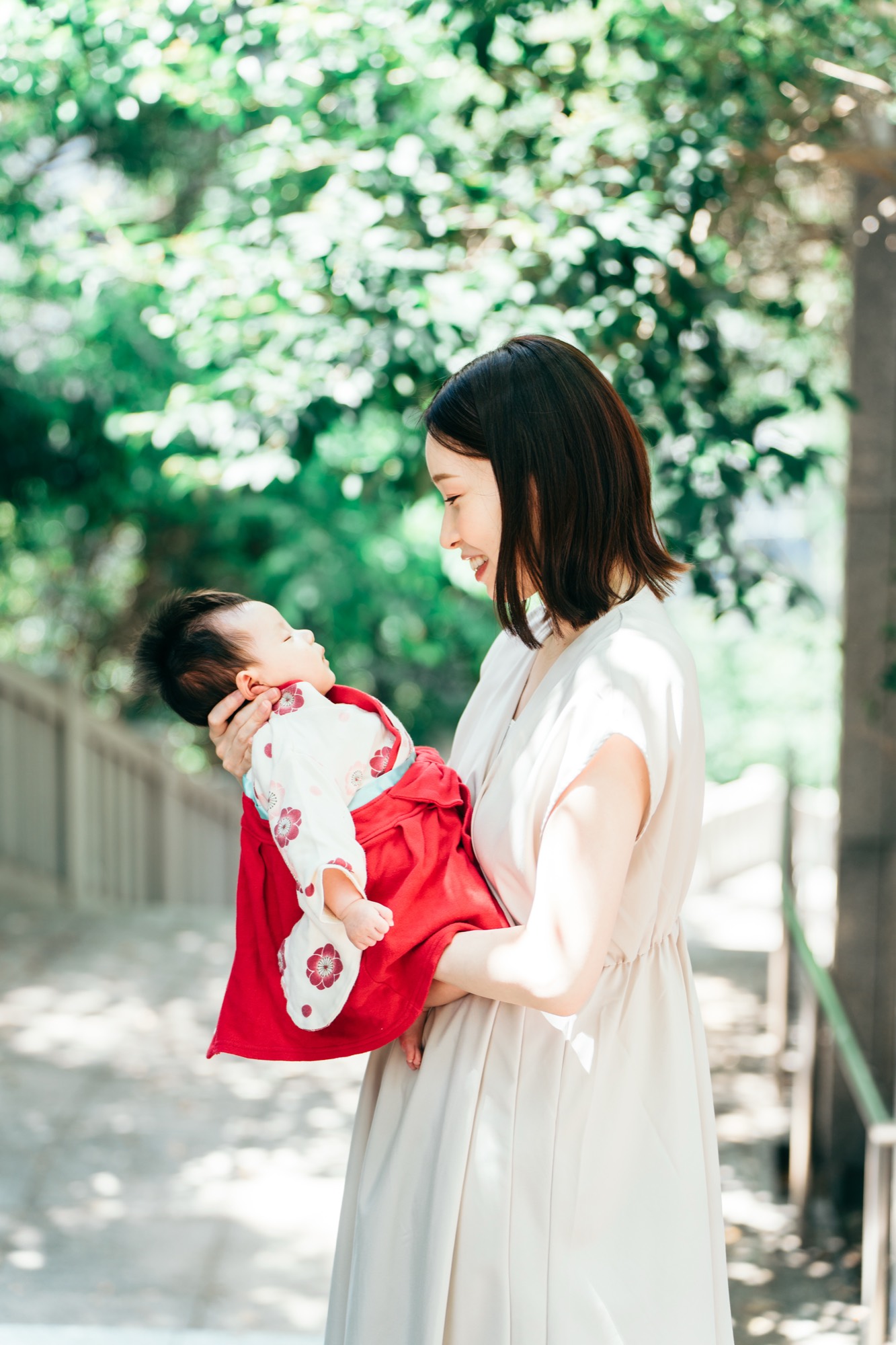 木漏れ日のなか、赤い祝い着の赤ちゃんをやさしく抱いて見つめるお宮参りのママ