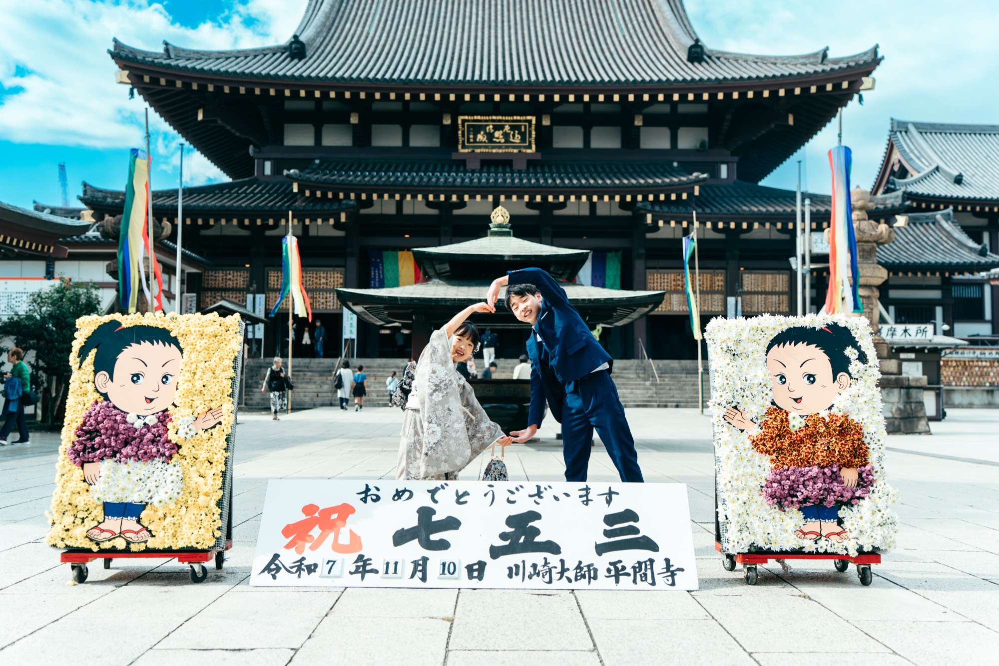 川崎大師の大本堂と「祝七五三」の花飾りを背景に、お父様とハートをつくる七五三の女の子