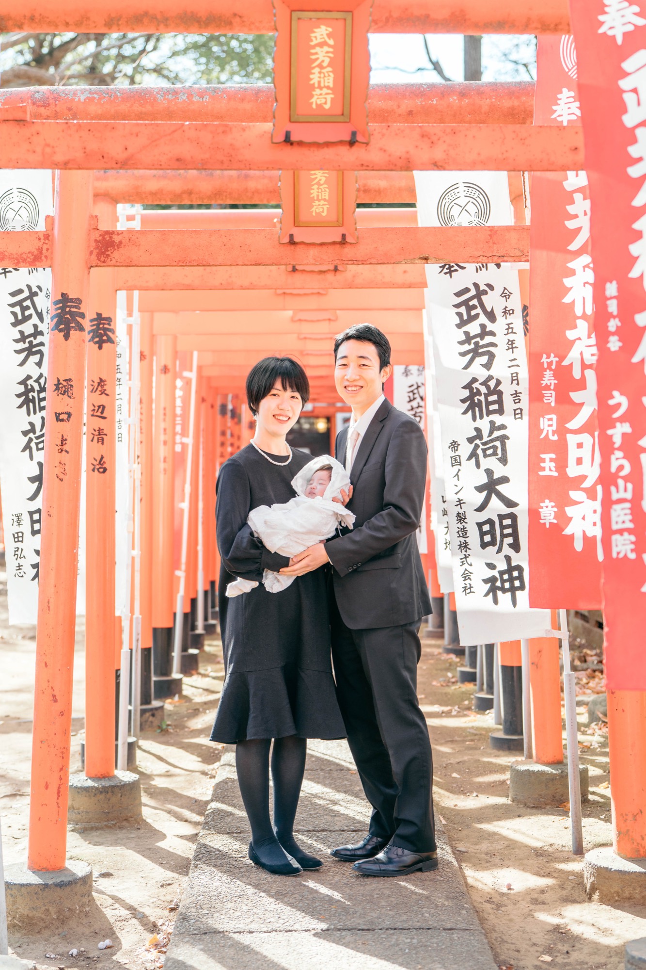 武芳稲荷の朱色の鳥居が連なるトンネルで、お宮参りの赤ちゃんを抱くご家族（雑司ヶ谷鬼子母神堂）