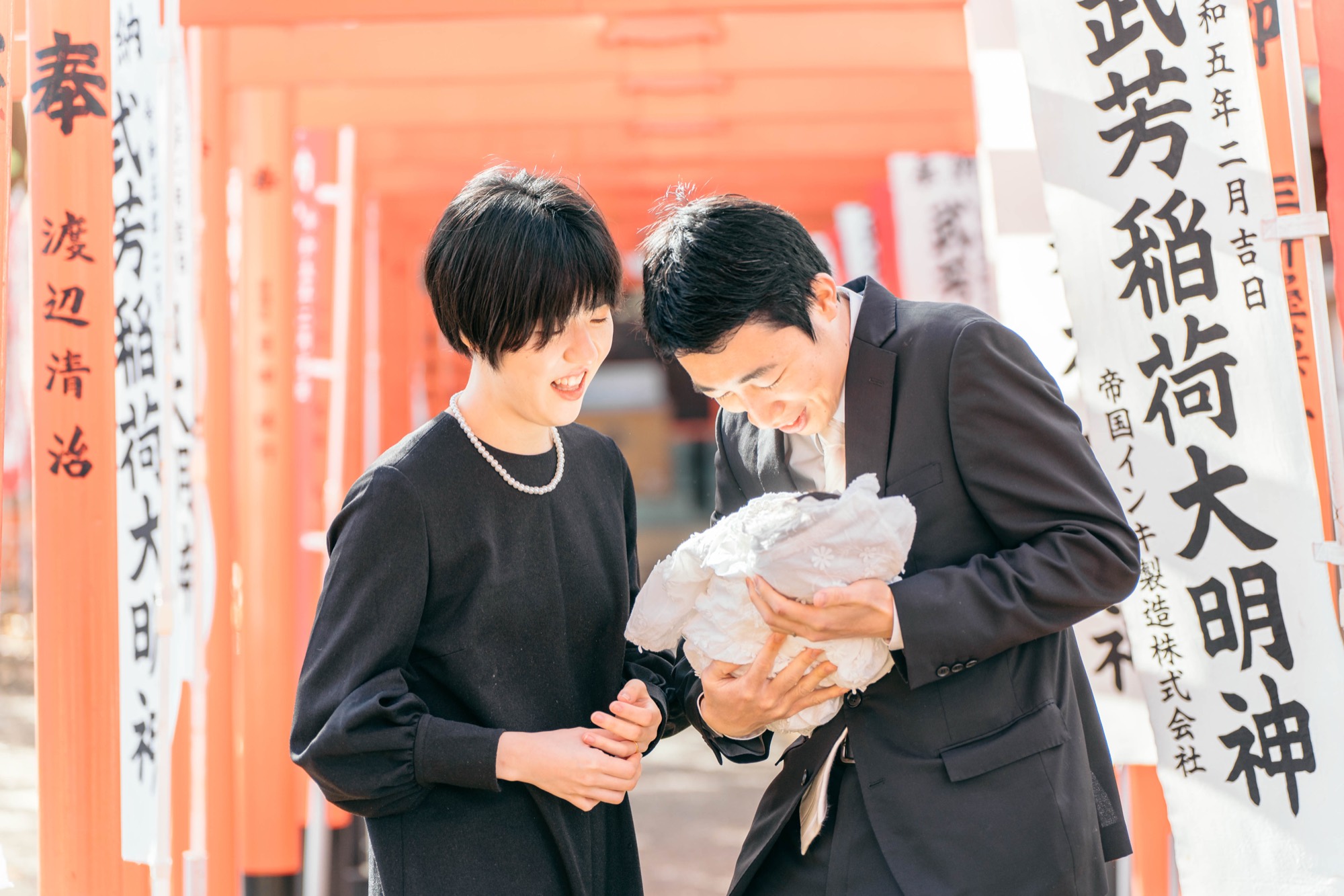 朱色の鳥居の前で祝い着の赤ちゃんを見つめて笑うご両親(鬼子母神堂・武芳稲荷)
