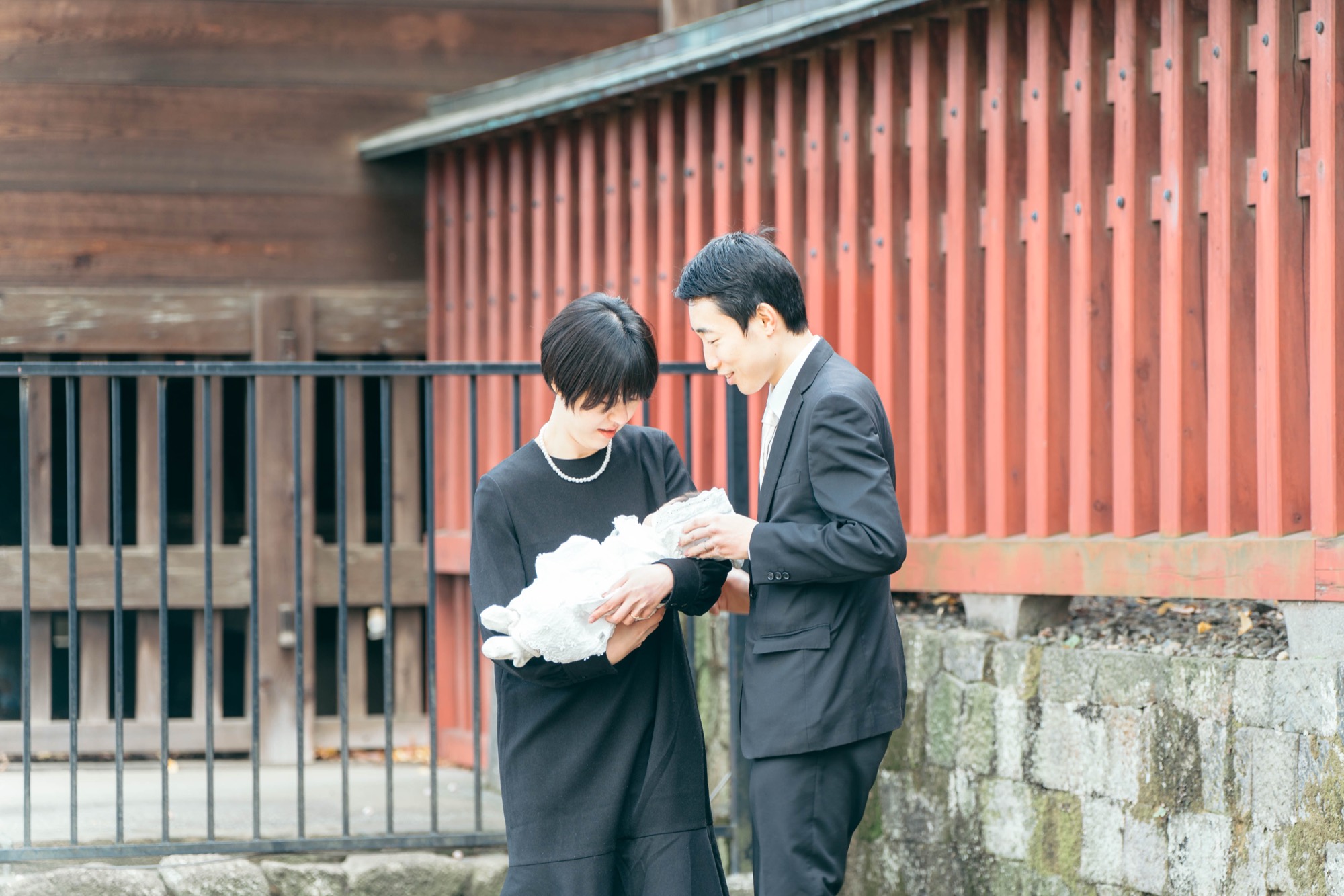 朱塗りの玉垣を背景に、白い祝い着の赤ちゃんをそっと抱くお宮参りのご家族