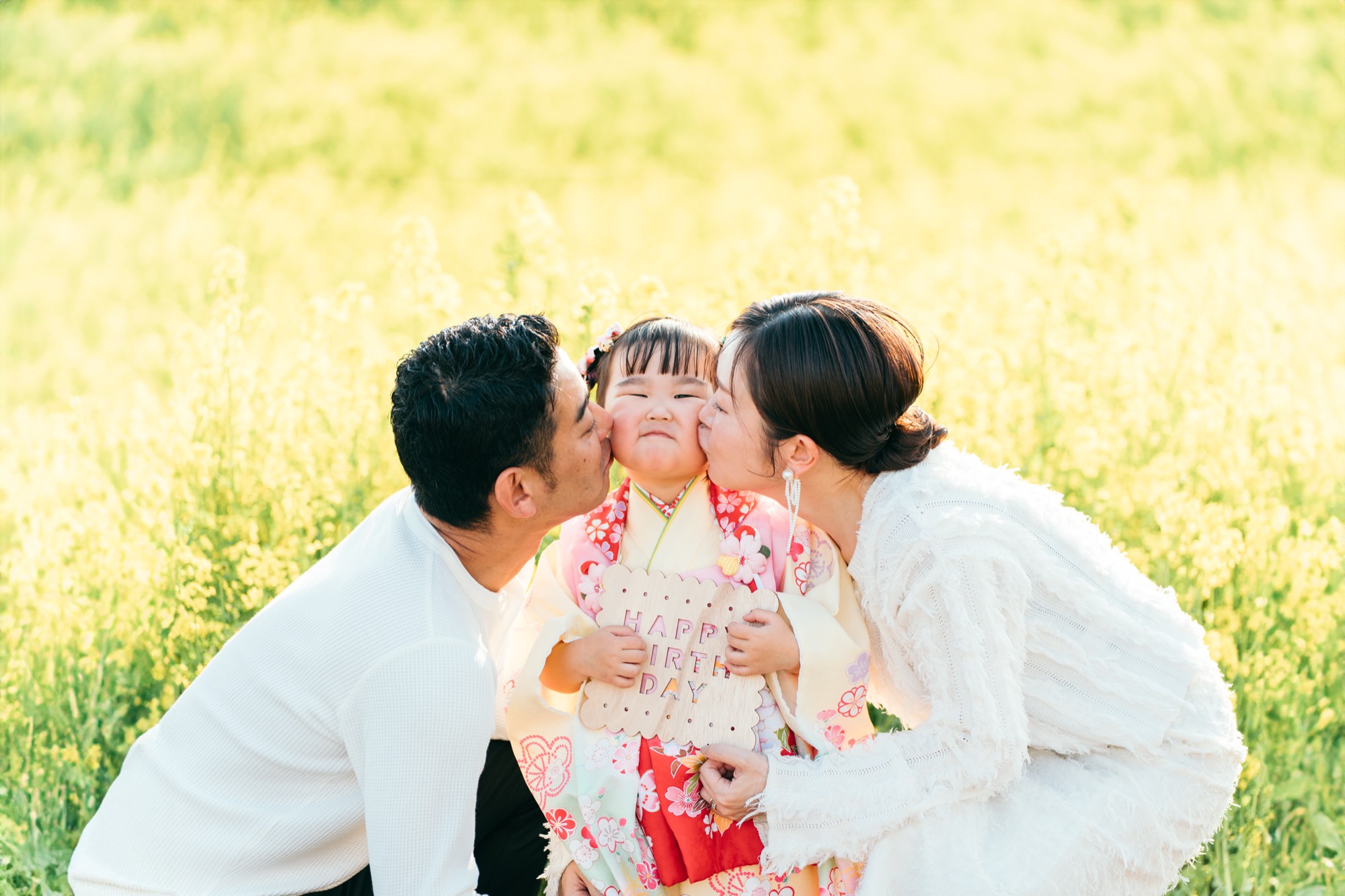 「HAPPY BIRTHDAY」のボードを持つ3歳の女の子の両頬に、パパとママがキスする菜の花畑の家族写真