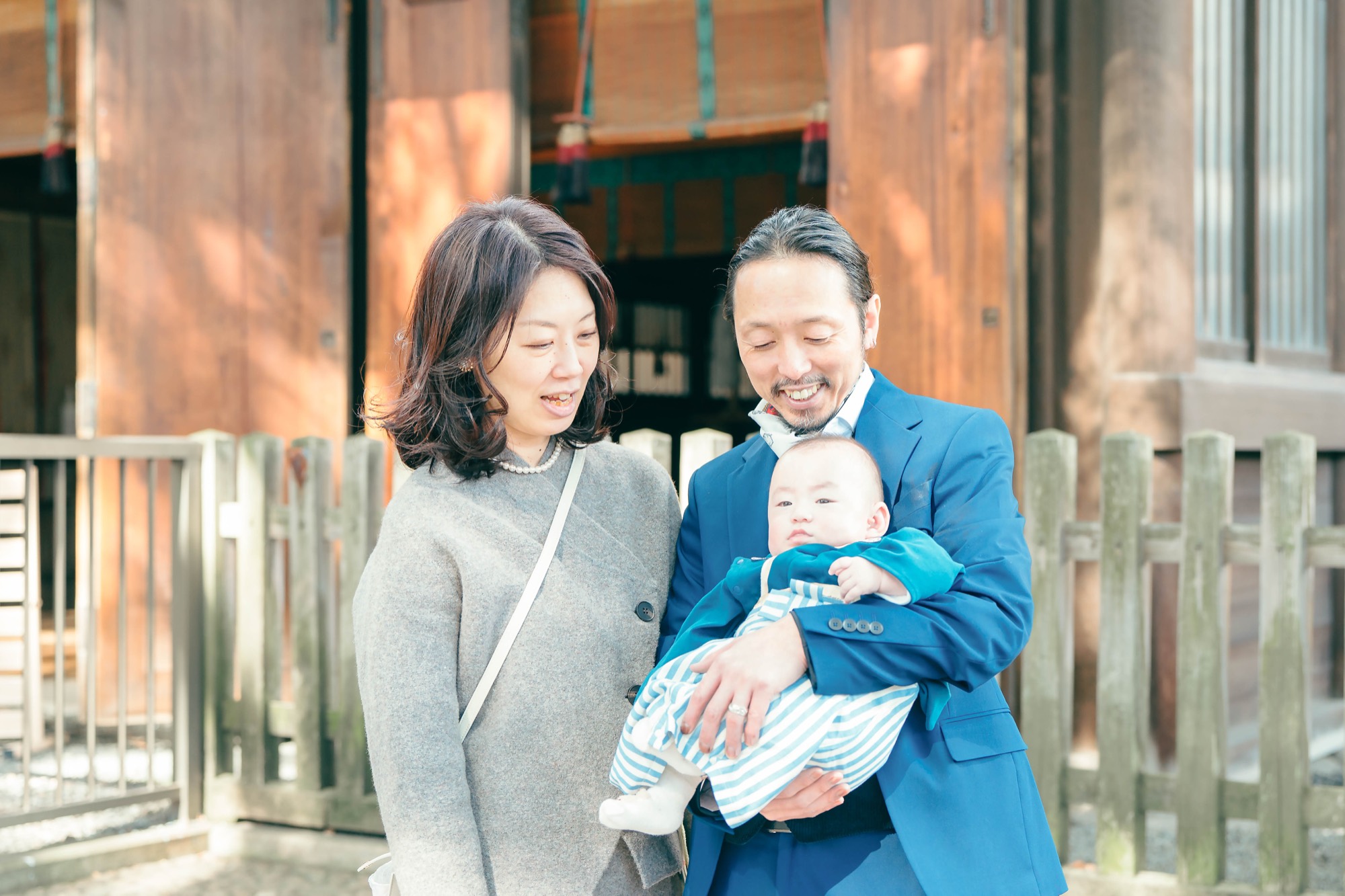 神社の境内で、お宮参りの祝い着姿の赤ちゃんを抱くパパとママの家族写真