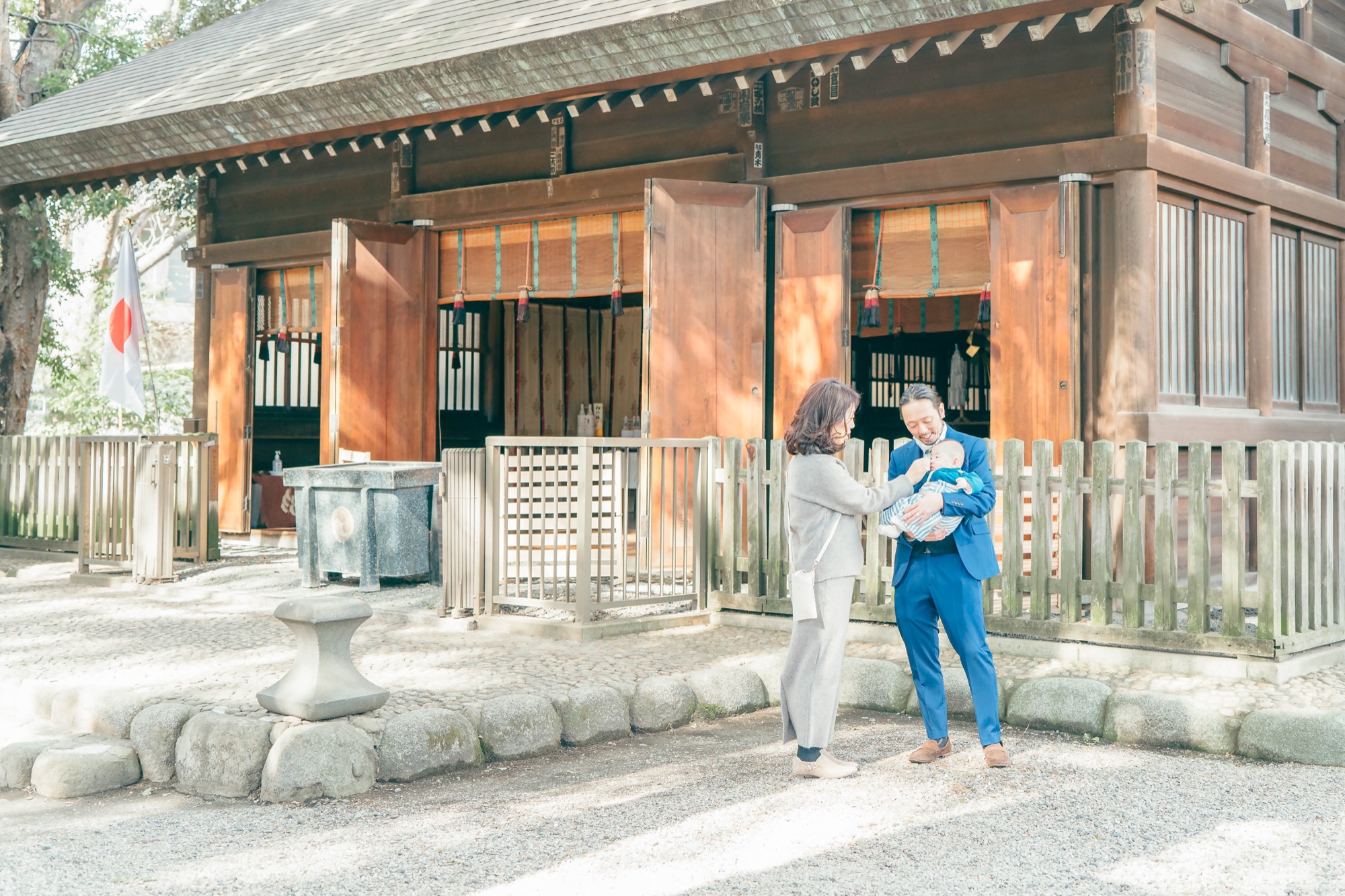 木造の社殿を背景に、赤ちゃんを囲んでお参りするお宮参りのご家族(祖父母も一緒に)