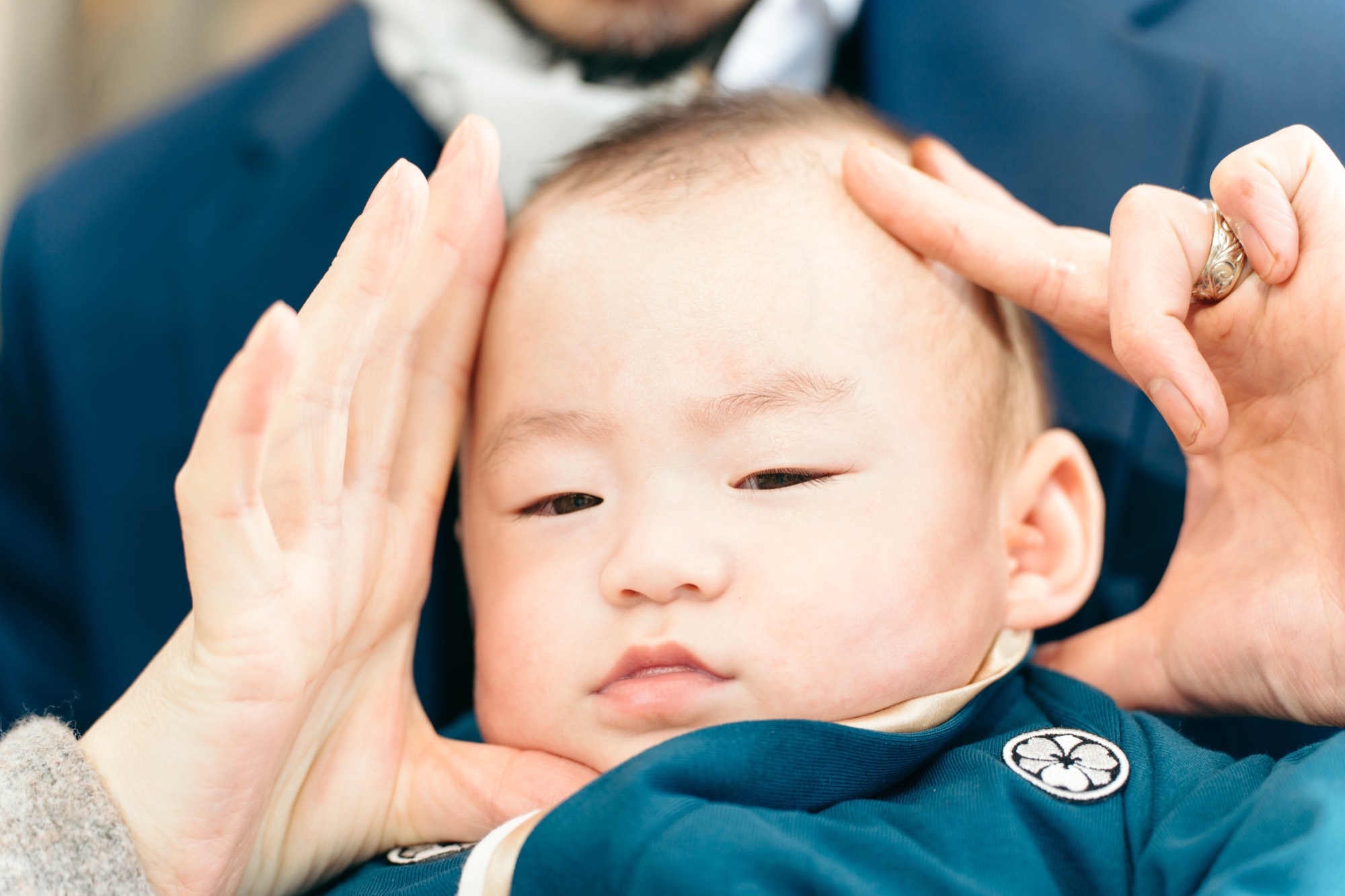 紺色の紋付き祝い着に包まれた、お宮参りの赤ちゃんのアップ。両親の手がそっと頬に添えられている