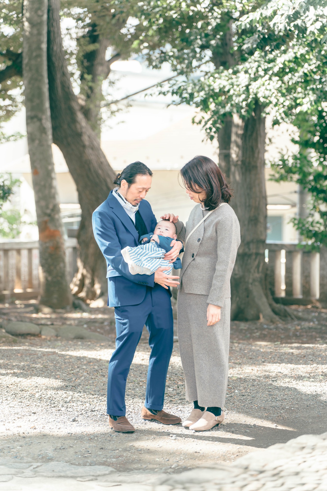落ち着いた境内の木陰で、祝い着の赤ちゃんをそっと抱いて見つめ合うお宮参りのご両親