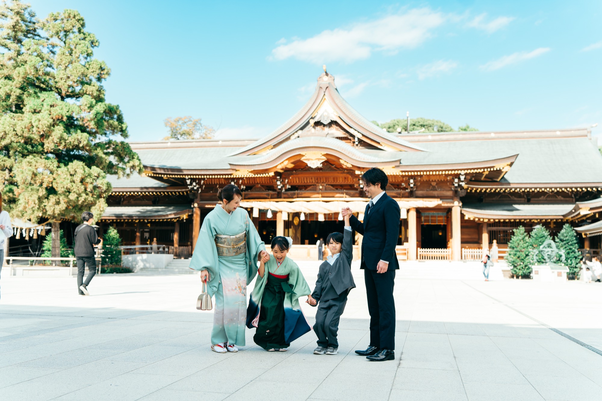 寒川神社の社殿を背景に、青空の下で手をつなぐ七五三の家族(袴姿の7歳の女の子とご家族)