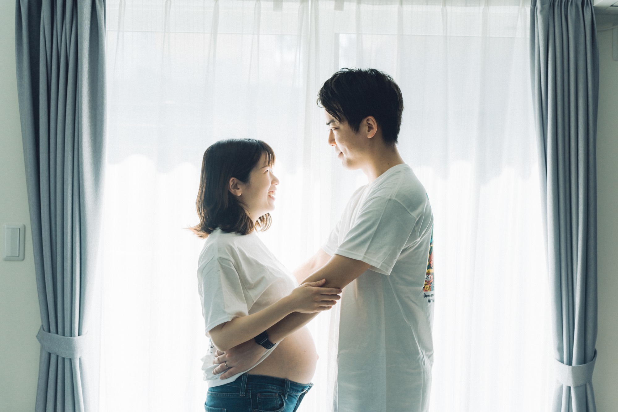 窓辺のやわらかな光のなか、おなかに手を添えて見つめ合うマタニティのご夫婦（ご自宅での出張撮影）