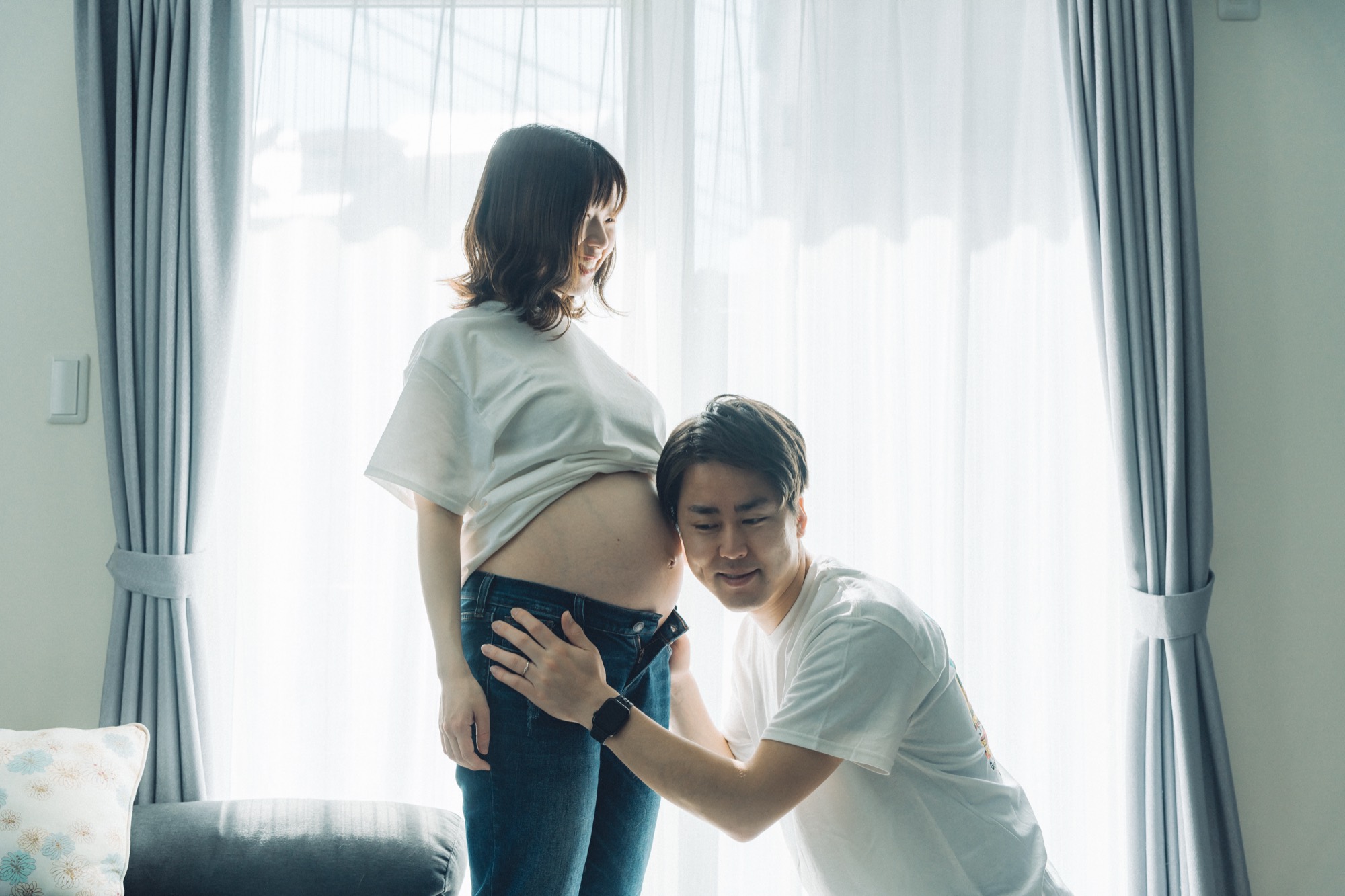 立つママのおなかにそっと耳を寄せるパパ。窓辺で撮影したマタニティフォト