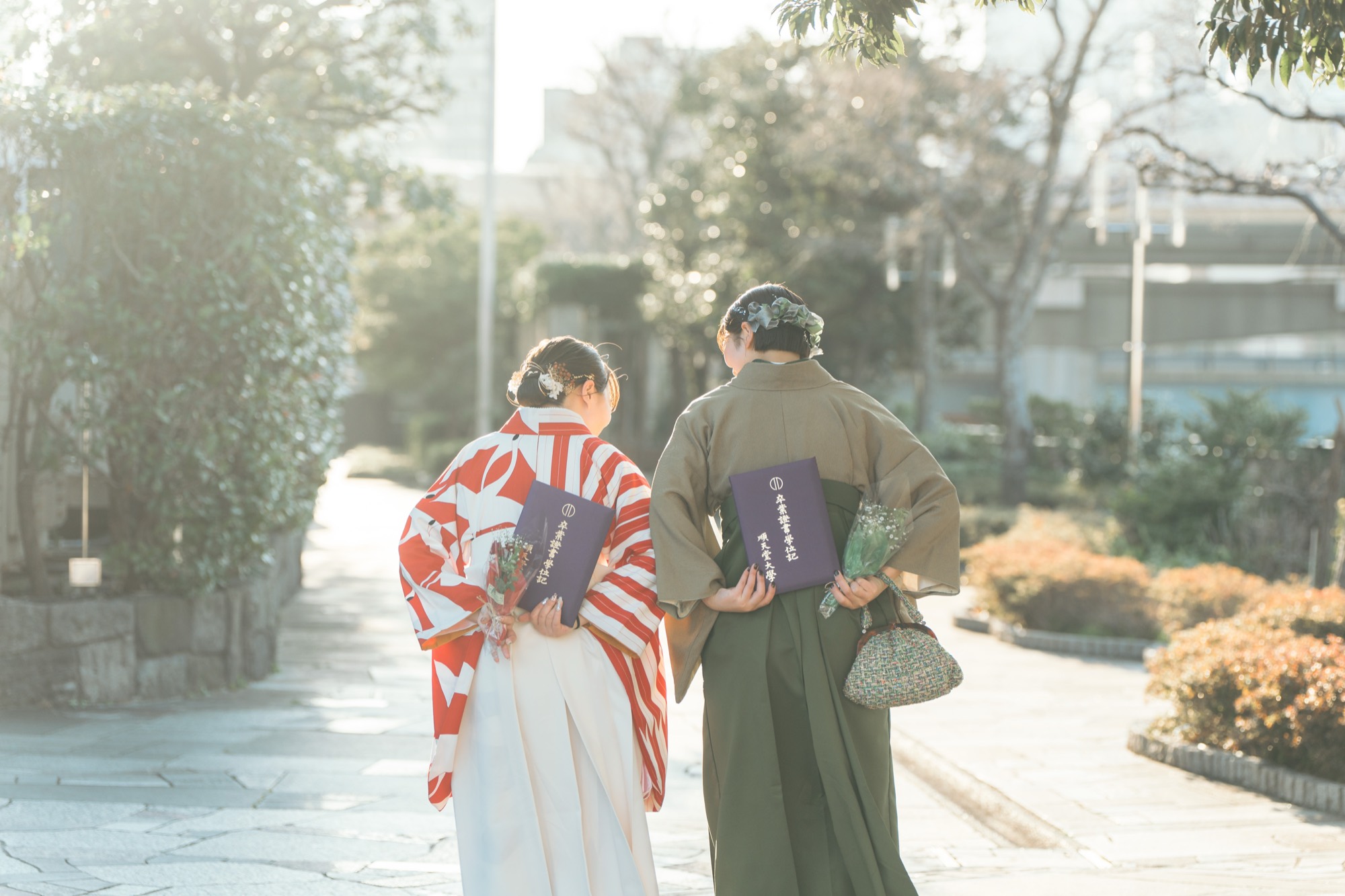 卒業証書と花束を手に並木道を歩く、袴姿の卒業生ふたりの後ろ姿